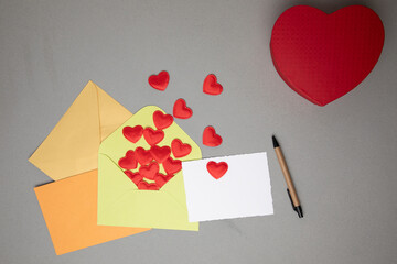 Light green, orange, yellow envelopes and a white card for text with red falling hearts on a gray concrete background. Happy Valentine's Day greeting, background. Flat lay