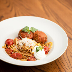 Portion of pasta with meatballs, sauce, tomatoes and cheese, garnished with basil leaves. Pasta in white plate on wooden table. Italian Cuisine. Side view. Copy space. Soft focus. 