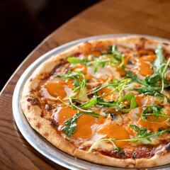 Italian pizza with cheese, ham, sauce and arugula baked intraditional way in oven. Ready dish on metal plate on wooden table. Dark background. View from above. Soft focus. Copy space. 