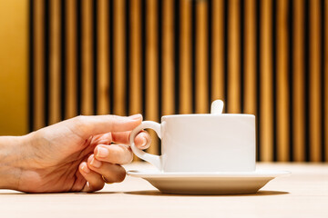 Hand of a young woman picking up white cup of coffee from the table