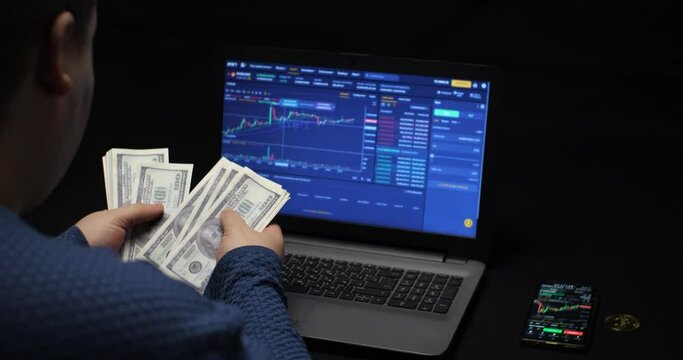 A trader counts dollars earned by selling cryptocurrencies on the stock exchange against the background of a graph on a laptop screen. A trader made money on bitcoin on a cryptocurrency exchange.