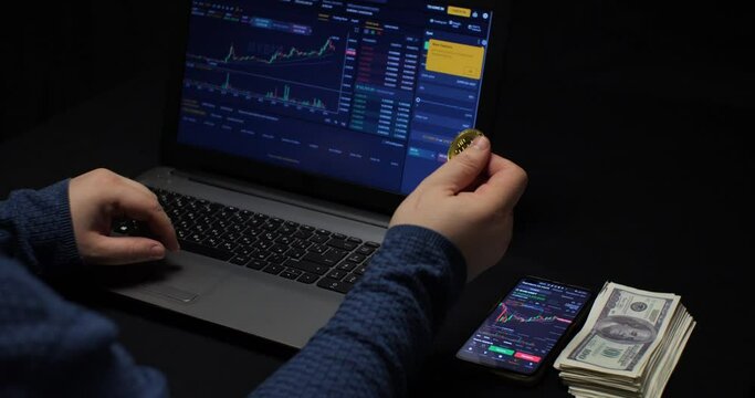 A man twists bitcoin in his hand while sitting at a laptop in the dark and watching the growth of the cryptocurrency chart on the stock exchange. Cryptocurrency mining on the electronic exchange.