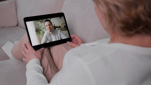An adult woman communicates with her son on a video call using a tablet while sitting in a cozy warm apartment on the couch. Living away from your parents. High quality 4k footage
