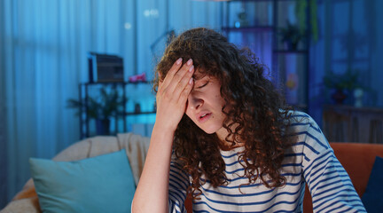 Displeased exhausted woman rubbing temples to cure headache problem, suffering from tension and migraine, stress, grimacing in pain, high blood pressure. Girl at home in evening room sitting on couch