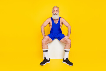 Fototapeta na wymiar Photo of sportive strong retired guy wear blue overall smiling sitting white cube chair isolated yellow color background