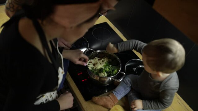 Family of little boy and young woman cooking in kitchen. Baby sitting near cooker, mixing soup with tablespoon in pan.