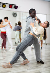 Fototapeta premium Smiling African American man supporting young girl, dancing slow foxtrot in pair in modern choreography studio