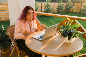 Laughing woman wearing pajama having video call on laptop during lunch break sitting at terrace in her home - summer time and video chat concept