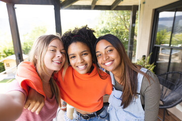 Portrait of happy diverse teenage female friends embracing on balcony taking selfie