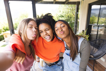 Portrait of happy diverse teenage female friends embracing on balcony taking selfie