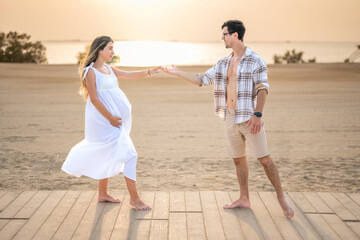 Love of pregnant woman and handsome man in unbuttoned shirt reaching each other hands at beach.