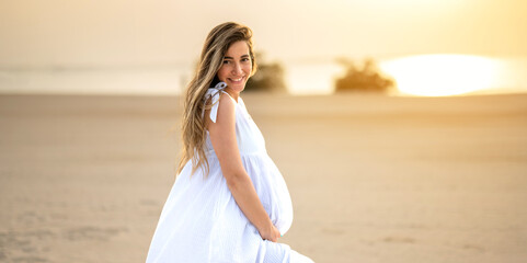 Profile view of pregnant woman with long hair in white dress on the beach at sunset