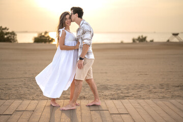 Fototapeta na wymiar Handsome man in a plaid shirt kissing his beautiful pregnant wife in a white dress at beach sunset.
