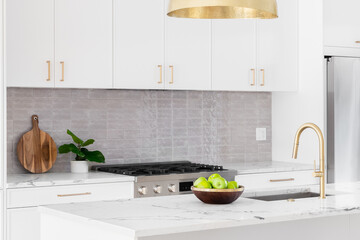 A kitchen detail with white cabinets, gold faucet and light hanging over the island, and a tiled backsplash.