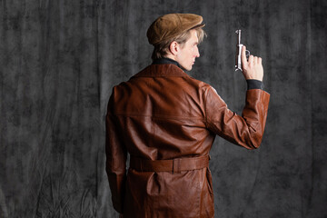 young man, a retro secret agent, a guy in a brown leather jacket and a flat cap with a gun in his hand. Posing in the studio on a gray background