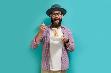 Portrait of a funny cute hipster guy with a beard in a hat opening a big mouth and eating noodles with a fork in the studio.