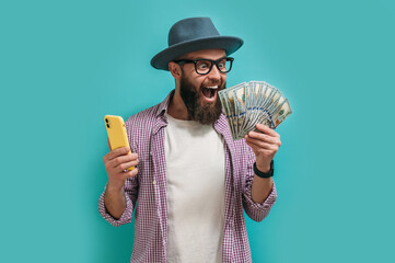 Excited happy young male winner feeling joy while holding money won, winning lottery, placing bets, getting cashback online gift on blue background. Human face emotions and betting concept. - obrazy, fototapety, plakaty