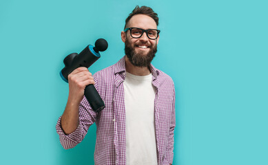Portrait of a young male physiotherapist in glasses for vision with a beard holding a massage gun in his hands isolated on blue studio background. Prevention of sports injuries. Health concept - obrazy, fototapety, plakaty