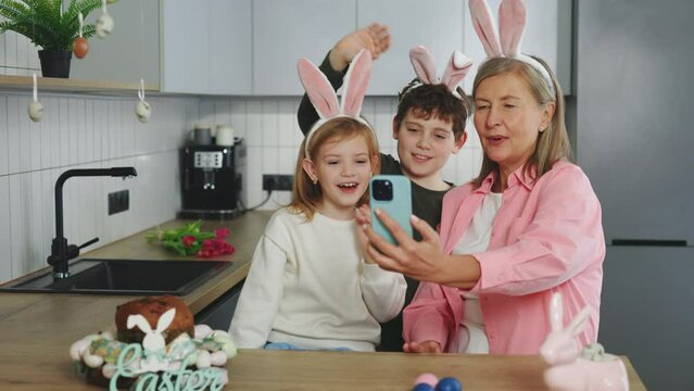 A Happy Grandmother and her Grandchildren are preparing for the Easter holiday using a Smartphone to video call. The family is painting eggs and communicating with friends through video. Happy Easter