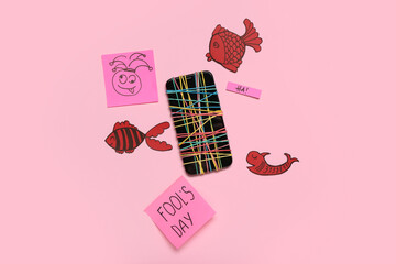 Mobile phone with rubber bands and paper decor on pink background. April Fool's Day celebration
