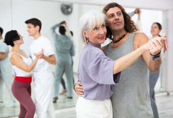 Fototapeta na wymiar Positive adult man and smiling aged woman practicing modern paired dance movements in group dance class