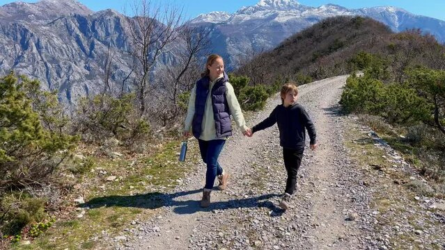 Young mother and son enjoy a scenic walk on a pebble pathway surrounded by majestic mountains, overlooking the stunning views of both Boka-Kotor Bay and Tivat Bay. It's the early spring or autumn in