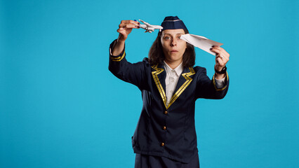 Fototapeta na wymiar Positive stewardess playing with toy and paper plane, having fun with origami and mini artificial airplane in studio. Happy air hostess wearing flying uniform working in aviation industry.