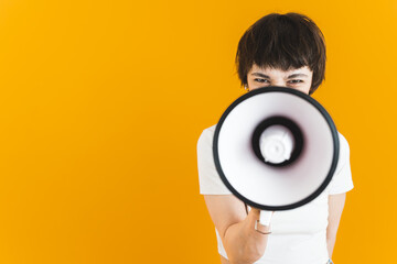Young angry protester shouting through loudspeaker. Frustrated woman frowning her eyebrows and holding megaphone in front of her face. Yellow background. High quality photo