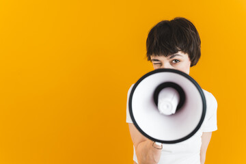 Young short-haired girl peeking behind white megaphone and blinking to the camera. Closeup front view studio portrait. Yellow background. High quality photo