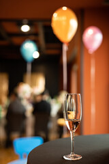 A glass of champagne is placed on a round black table. In the background are people participating in the party and colored helium balloons