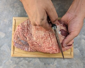 Pair of hands cutting across picaña meat lard with an iron knife on wooden plate seen from top