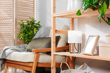 Wooden shelving unit with home decor, folding screen and armchair near light wall in room. Interior design