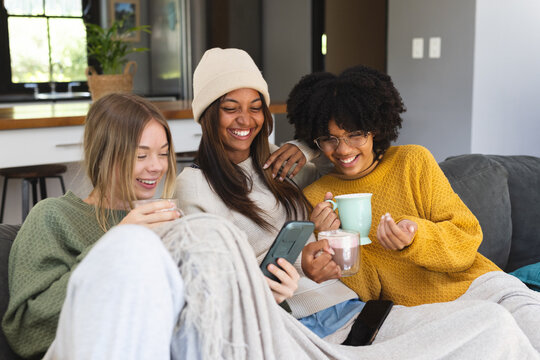 Happy diverse teenage female friends using smartphone and drinking cocoa