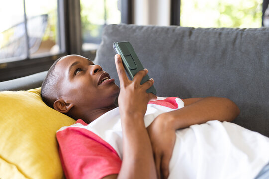 Happy african american teenage boy lying on sofa and using smartphone