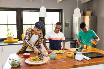 Happy diverse teenage male friends cutting self made pizza in kitchen