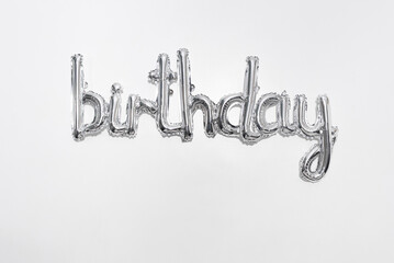 Silver birthday balloon lettering center on a white wall. Foil letters isolated on horizontal background. Metallic word, party decor, shiny typography.