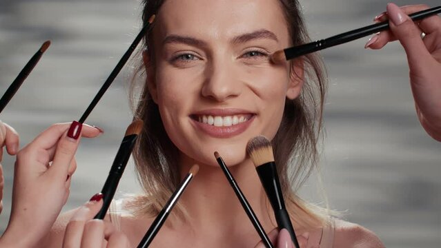 Team of specialists giving makeover to beautiful girl, make up artists using brushes to create glowing make up look on woman. Smiling confident lady receiving nourishing healthy treatment. Close up.