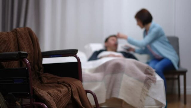Close-up wheelchair indoors with blurred woman feeding man lying in bed at background. Unrecognizable Caucasian wife taking care of sick husband at home. Lifestyle and love concept