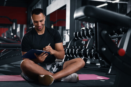 Happy trainer writing down workout plan in modern gym, space for text