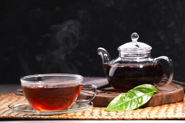 Aromatic hot tea in glass cup with saucer, teapot and leaves on table against black background. Space for text