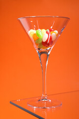 Martini glass with many tasty colorful candies on table against orange background