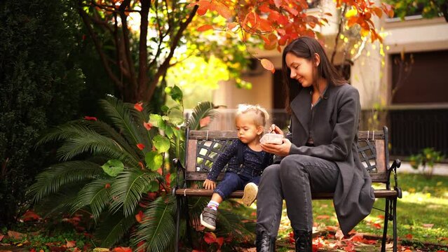 Mom feeds a little girl with a spoon sitting on a bench in the garden with colorful leaves