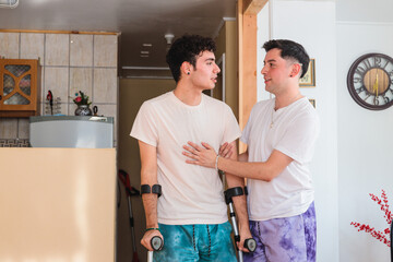 Homosexual man supporting his injured partner who is using crutches inside their house. LGBT...