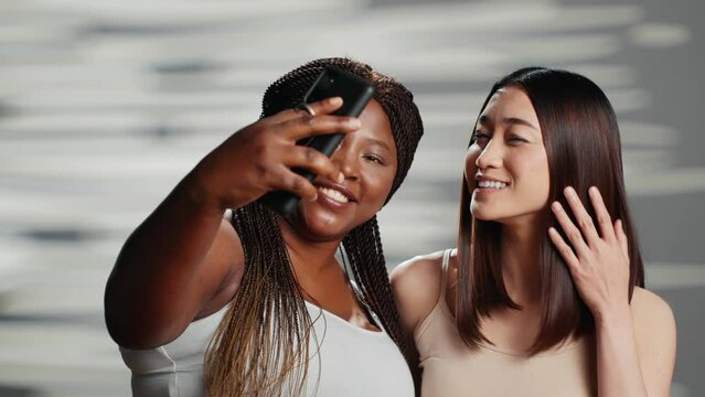 Diverse friends taking photos on mobile phone in studio, expressing self love and self confidence. Young beauty models promoting body positivity and different skintones, wellness.