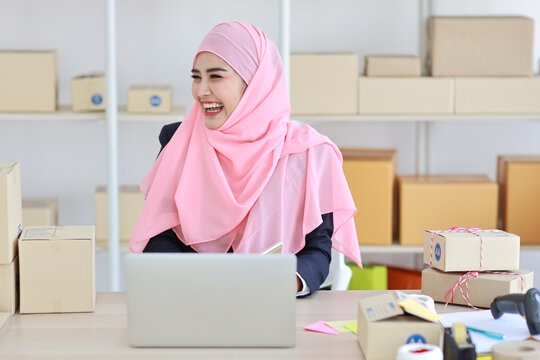 Religious Asian Woman In Muslim Dress Sitting And Using Smart Mobile Phone And Talking To Customer With Happy Face And Business Package SME Box Delivery Background.