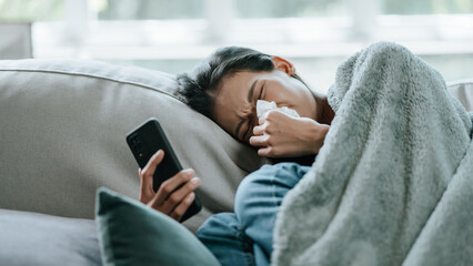 Asian woman in cloak suffering from fever and flu on sofa while using mobile phone