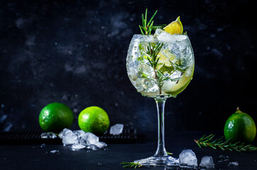 Gin tonic cocktail with dry gin, rosemary, tonic, lime and ice cubes in wine glass. Black bar counter background, bar tools, copy space - obrazy, fototapety, plakaty