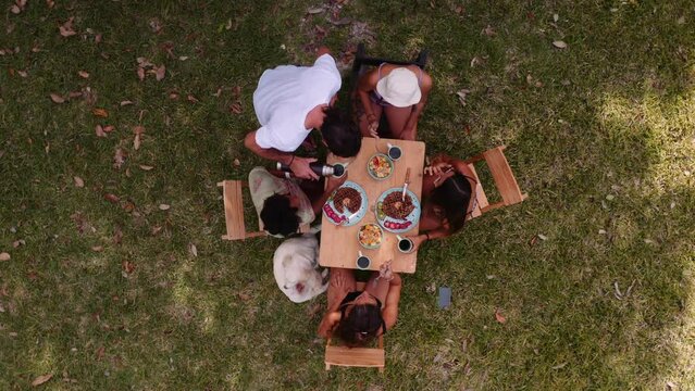 Aerial top down drone shot of man serving coffee to four women sitting around a wooden table and eating hot cakes. Sunny day, in the grass.
