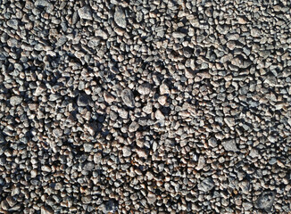 Small dark grey granite stones lie on a dirt road, top view in the rays of the midday sun.
