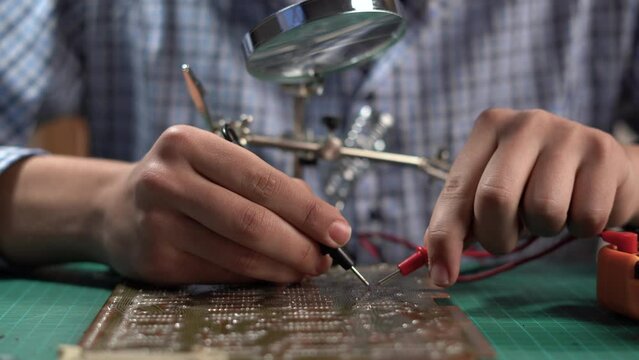 Teen boy using a test head connected to the multimeter while examining a circuit board placed under the magnifying glass. Teenager hobbies concept.
